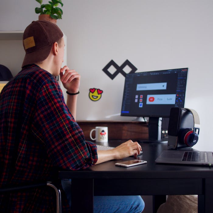 guy weaaring a haat and a shiirt looking at computer monitor, by using a macbook