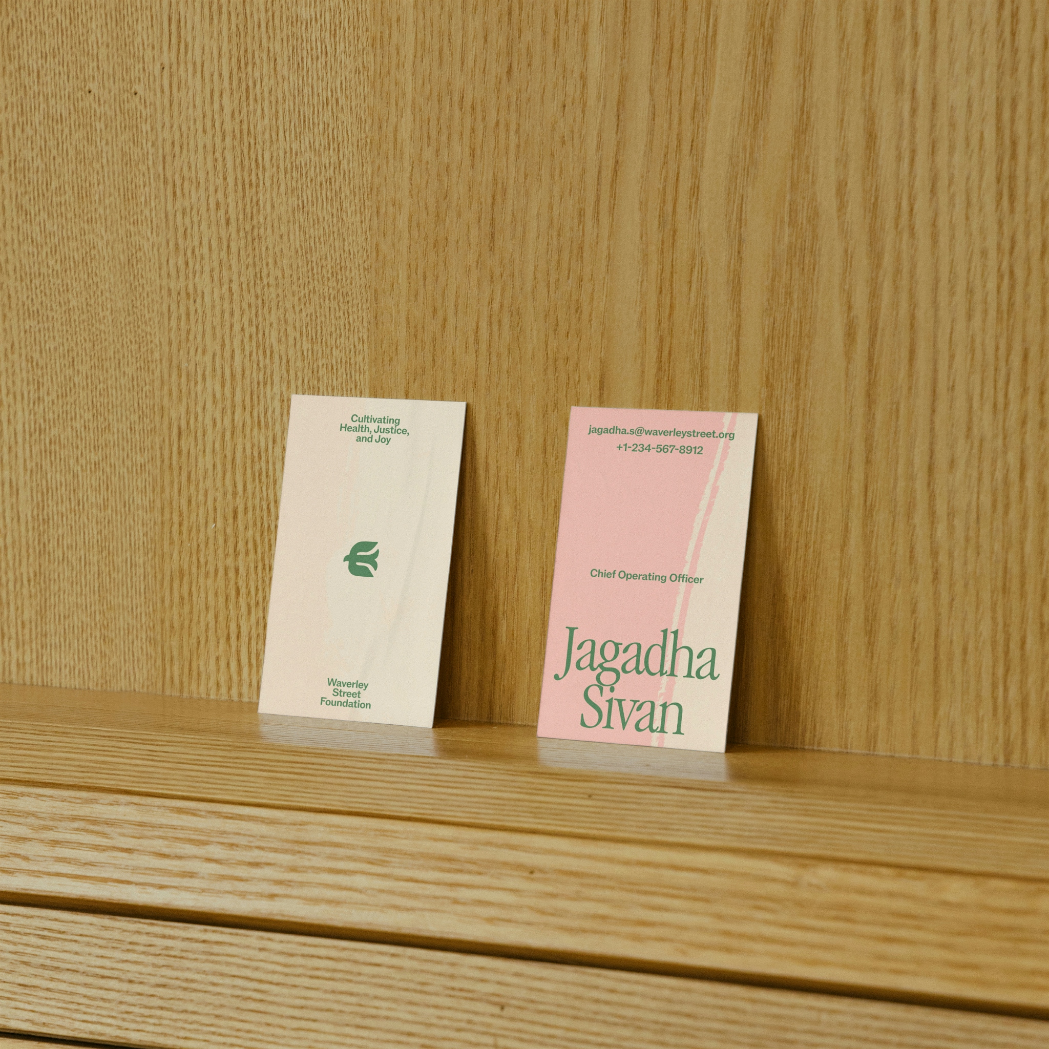 Two business cards for Waverley Street Foundation displayed on a light wood shelf. The left card is cream-colored with green text reading 'Cultivating Health, Justice, and Joy' and features the foundation's logo. The right card is pink with green text identifying 'Jagadha Sivan, Chief Operating Officer' along with her email address and phone number.