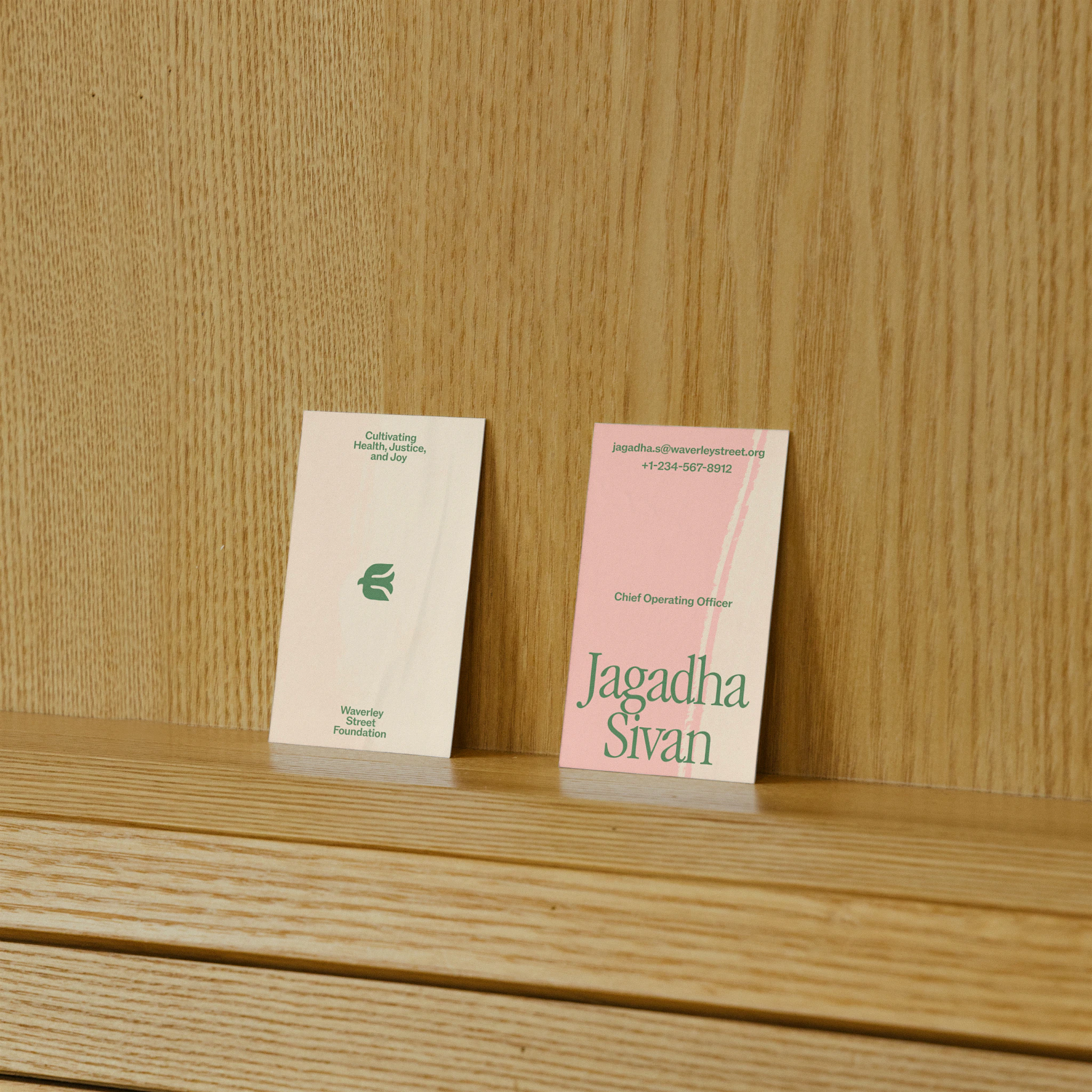 Two business cards for Waverley Street Foundation displayed on a light wood shelf. The left card is cream-colored with green text reading 'Cultivating Health, Justice, and Joy' and features the foundation's logo. The right card is pink with green text identifying 'Jagadha Sivan, Chief Operating Officer' along with her email address and phone number.