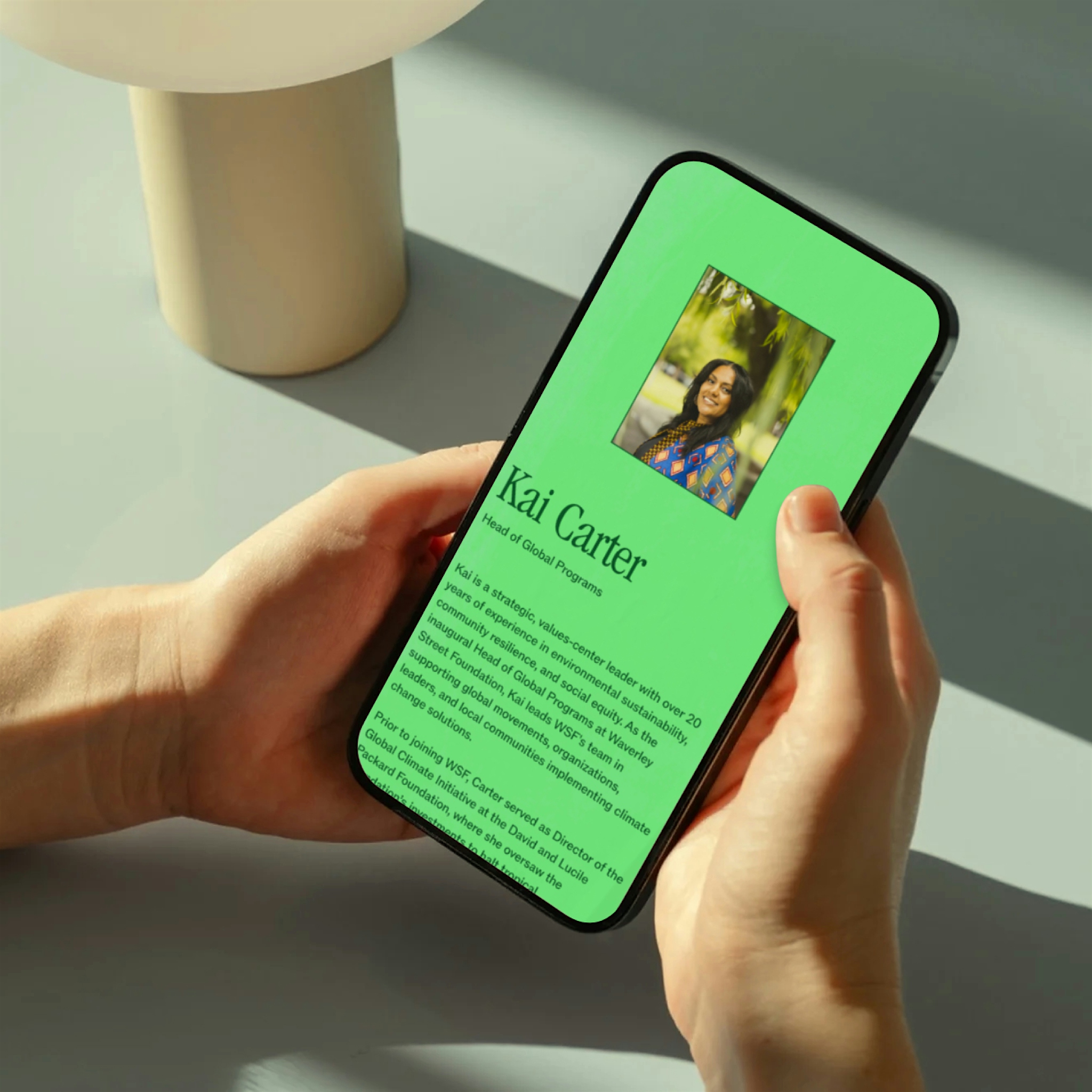 Hands holding a smartphone displaying a bright green staff profile for Kai Carter, Head of Global Programs at Waverley Street Foundation, featuring a photo and brief professional biography highlighting environmental leadership experience.