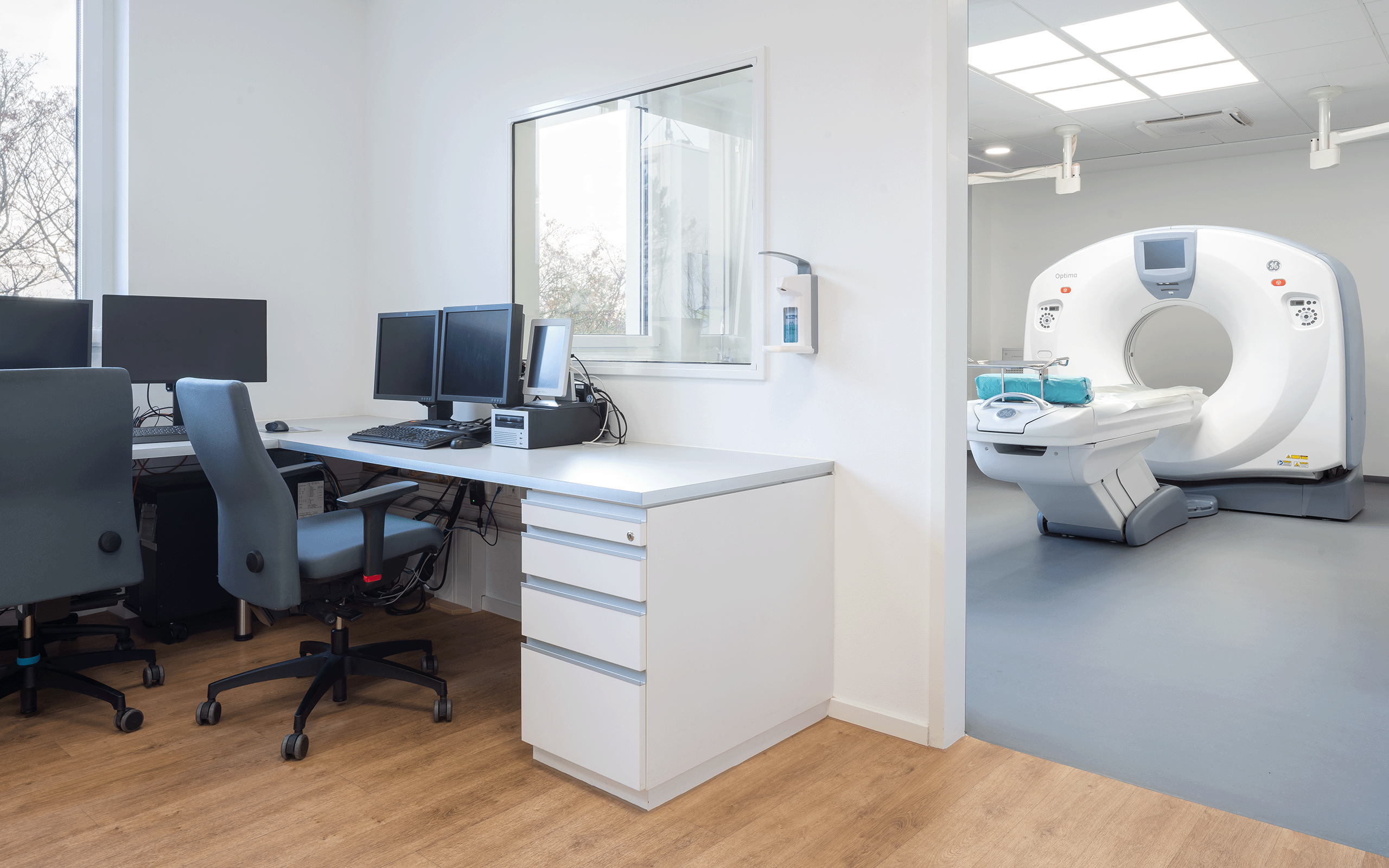 Praxiszimmer mit Schreibtisch und CT-Gerät im Nebenraum