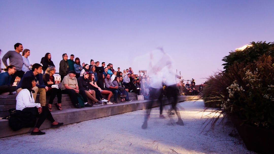 People sit on the wooden platforms at dusk at the Rail Park. Blurry figures of people dancing are seen along the path.