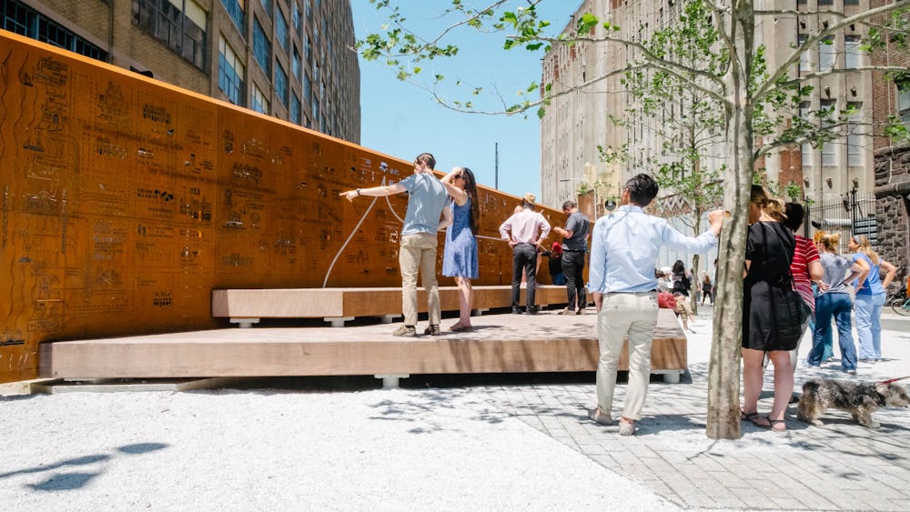 A group of people stand in front of the Story Wall, an 80 foot galvanized steel wall with a historic atlas map, at the entrance of Phase One.