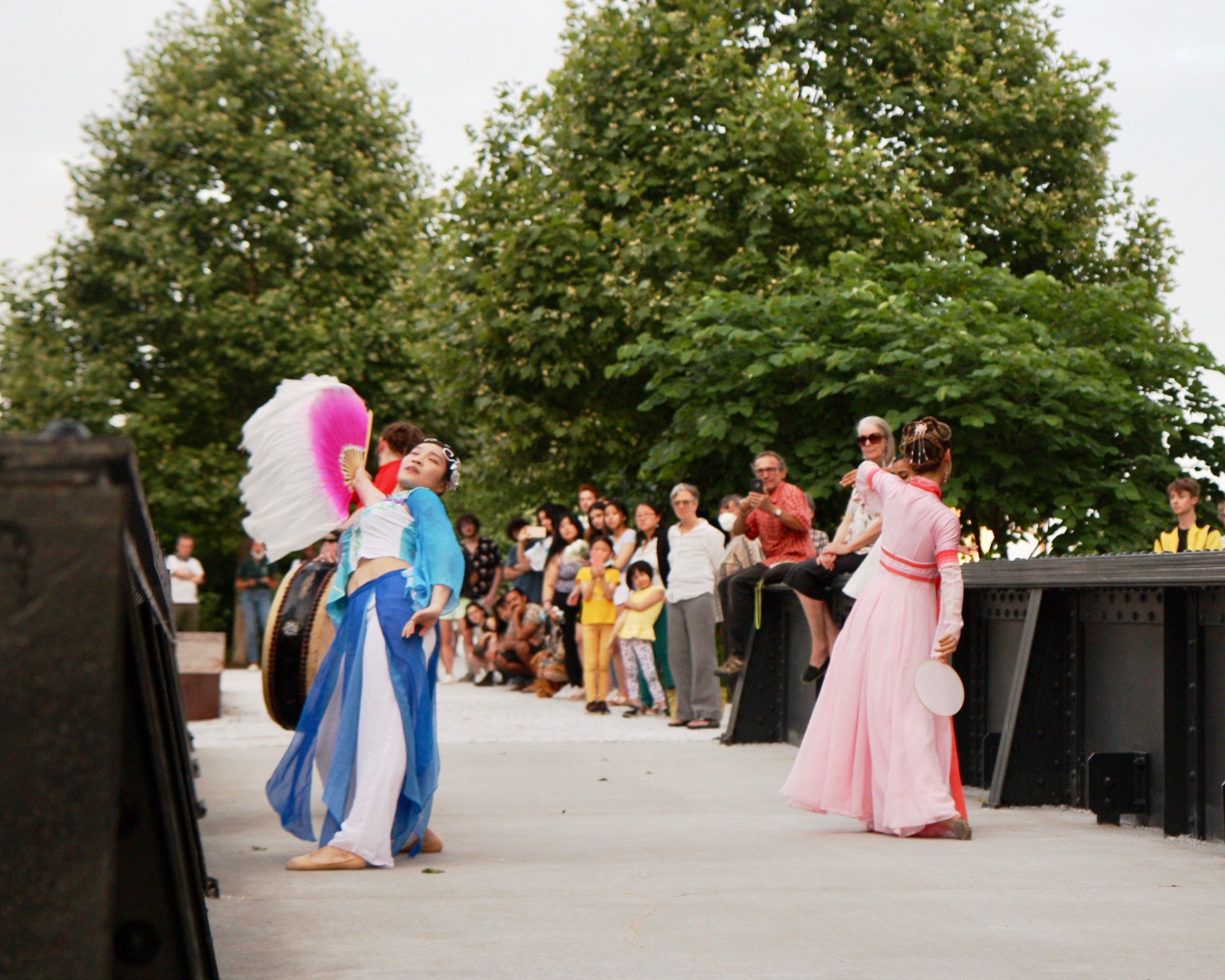 Three performers, one wearing a pink and red dress and the other wearing a blue and white dress, and the other playing a drum, dance towards a crowd of people across a train bridge at the Rail Park.