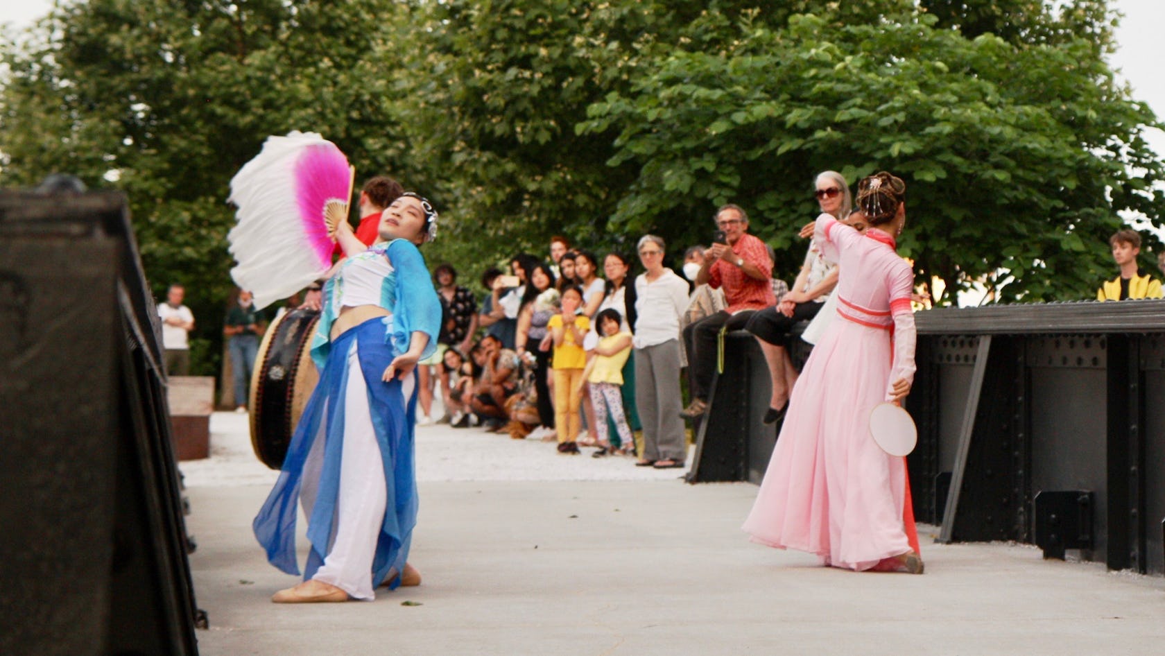 Three performers, one wearing a pink and red dress and the other wearing a blue and white dress, and the other playing a drum, dance towards a crowd of people across a train bridge at the Rail Park.