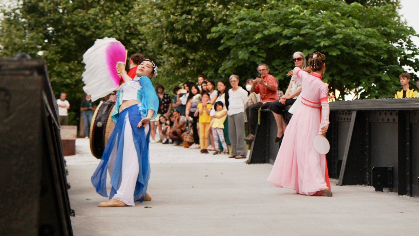 Three performers, one wearing a pink and red dress and the other wearing a blue and white dress, and the other playing a drum, dance towards a crowd of people across a train bridge at the Rail Park.