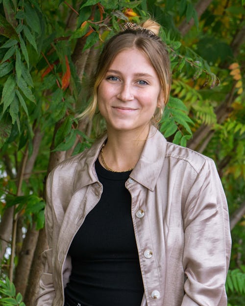 Professional headshot of Ava Schwemler. A person with blond hair and light skin that is pulled back into a bun, with some strands of hair framing her face. She is wearing a black shirt and a gold button down that is open in the front. She is smiling and standing against a background of sumac trees at the Rail Park. 