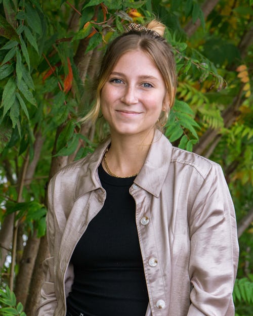 A person with blond hair and light skin that is pulled back into a bun, with some strands of hair framing her face. She is wearing a black shirt and a gold button down that is open in the front. She is smiling and standing against a background of sumac trees at the Rail Park.