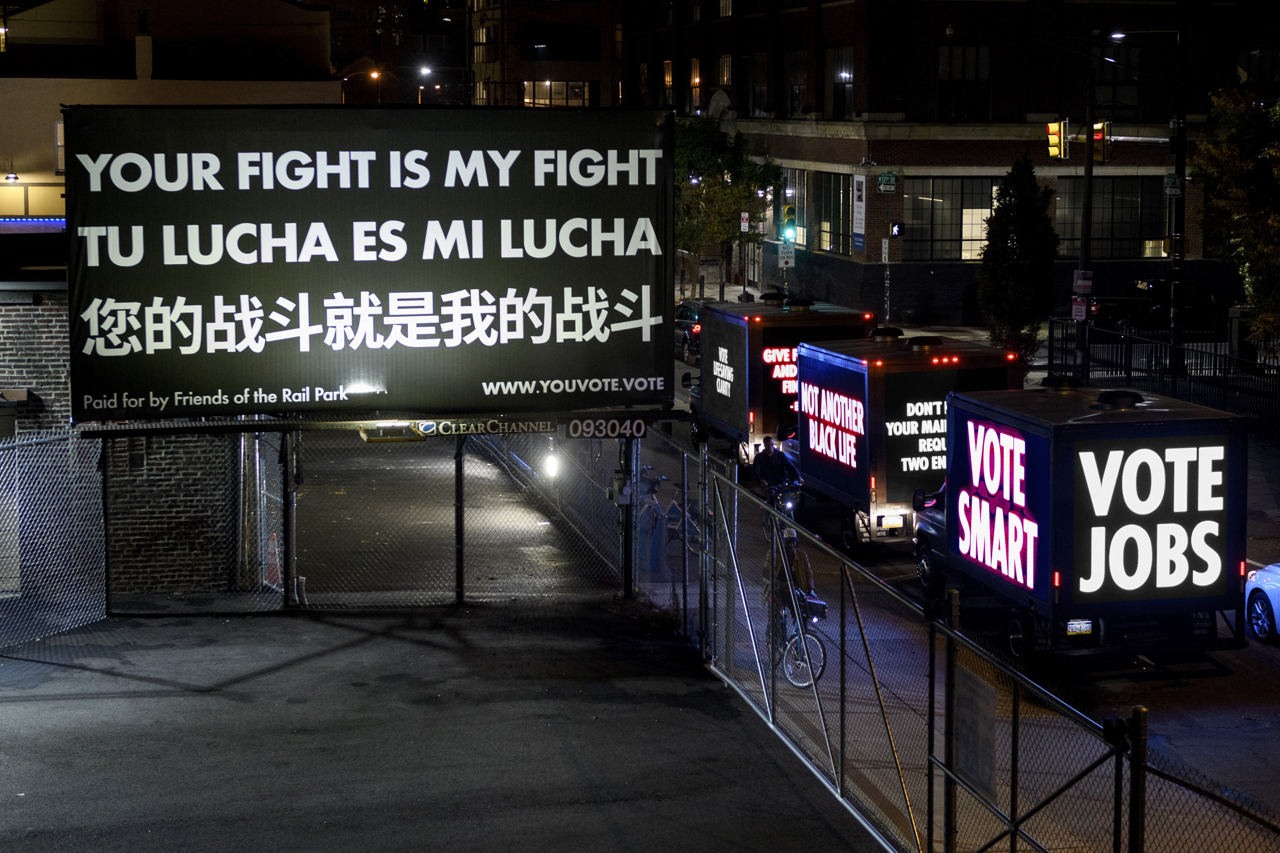 At nighttime, lights illuminate a black and white billboard that reads "YOUR FIGHT IS MY FIGHT" in English, Spanish, and Mandarin. 3 Trucks with LED screens reading "VOTE JOBS", "VOTE SMART" drive by the street below. 