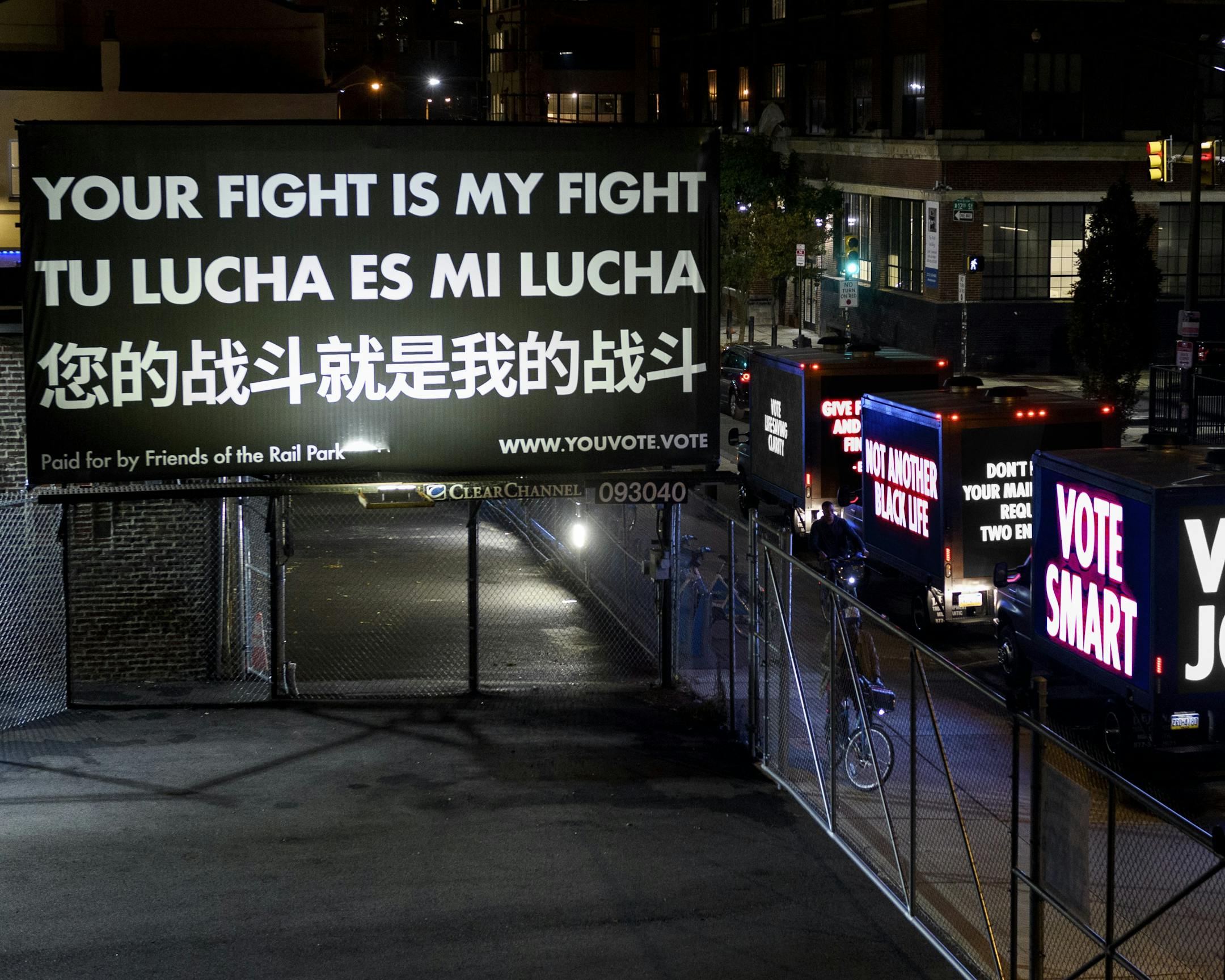At nighttime, lights illuminate a black and white billboard that reads "YOUR FIGHT IS MY FIGHT" in English, Spanish, and Mandarin. 3 Trucks with LED screens reading "VOTE JOBS", "VOTE SMART" drive by the street below. 