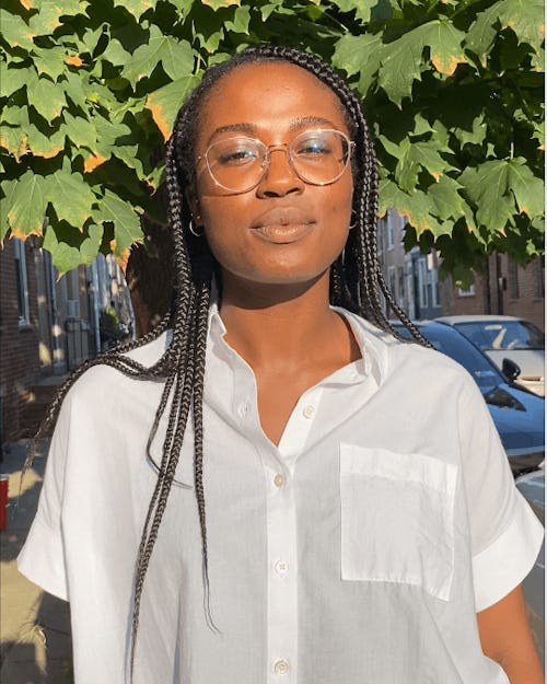 A person with long, dark, braided hair wears glasses and a white shirt. They wear glasses and have a neutral expression. Behind them, the leaves of a tree are visible