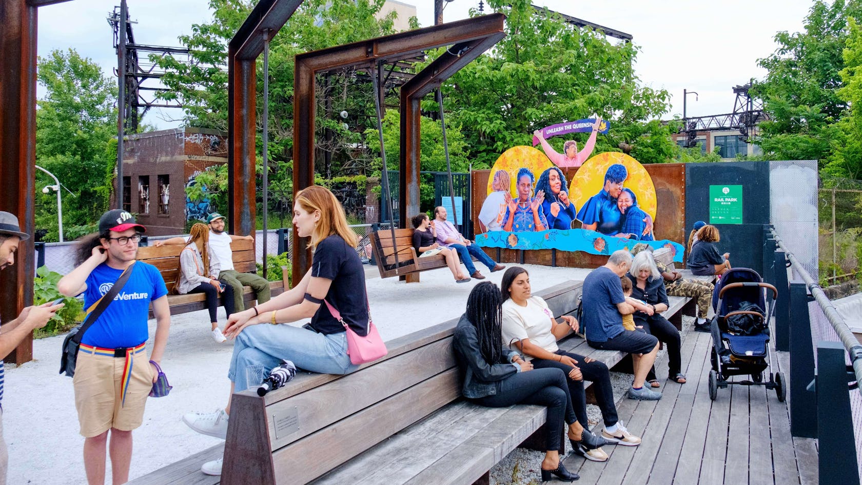 Photo shows a scene at the Rail Park with many people hanging out, talking to each other, relaxing, etc. In the background, a colorful and joyful artwork is visible, displaying portraits of 6 women