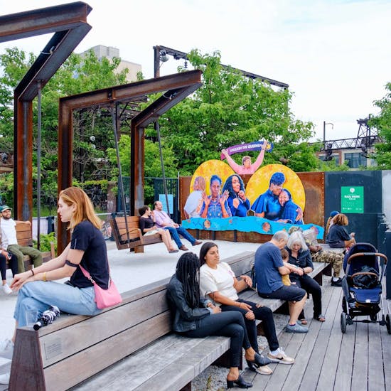 Photo shows a scene at the Rail Park with many people hanging out, talking to each other, relaxing, etc. In the background, a colorful and joyful artwork is visible, displaying portraits of 6 women