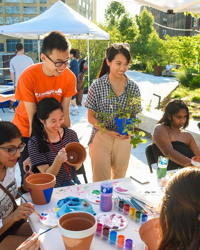 A group of people surrounds a table filled with pots and paints. People are painting the pots and a person holds plants to put into the pots. 
