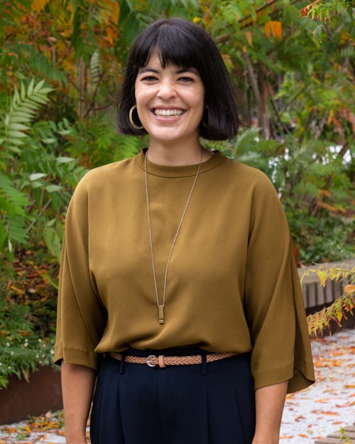 A woman with dark brown hair that is cut into a bob. She is wearing dark blue pants and a brown shirt. She is smiling and showing her teeth. She is standing with her hands at her sides at the Rail Park with sumac trees in the background. 