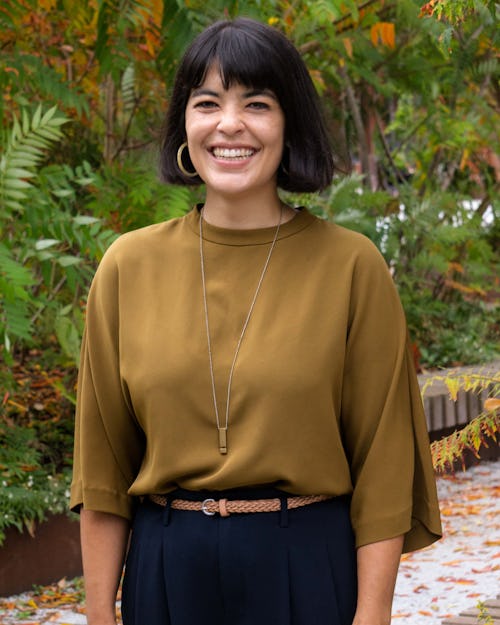 Person with dark brown hair that is cut into a bob. She is wearing dark blue pants and a brown shirt. She is smiling and showing her teeth. She is standing with her hands at her sides at the Rail Park with sumac trees in the background. 