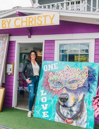 Artist Christy Boutte standing with her painting of Raising Cane 3 in a Mardi Gras feather mask.