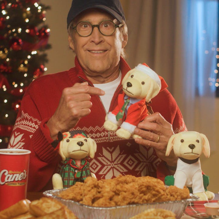 Chevy Chase holding a holiday Plush Puppy sitting in front of a large tray of Chicken Fingers