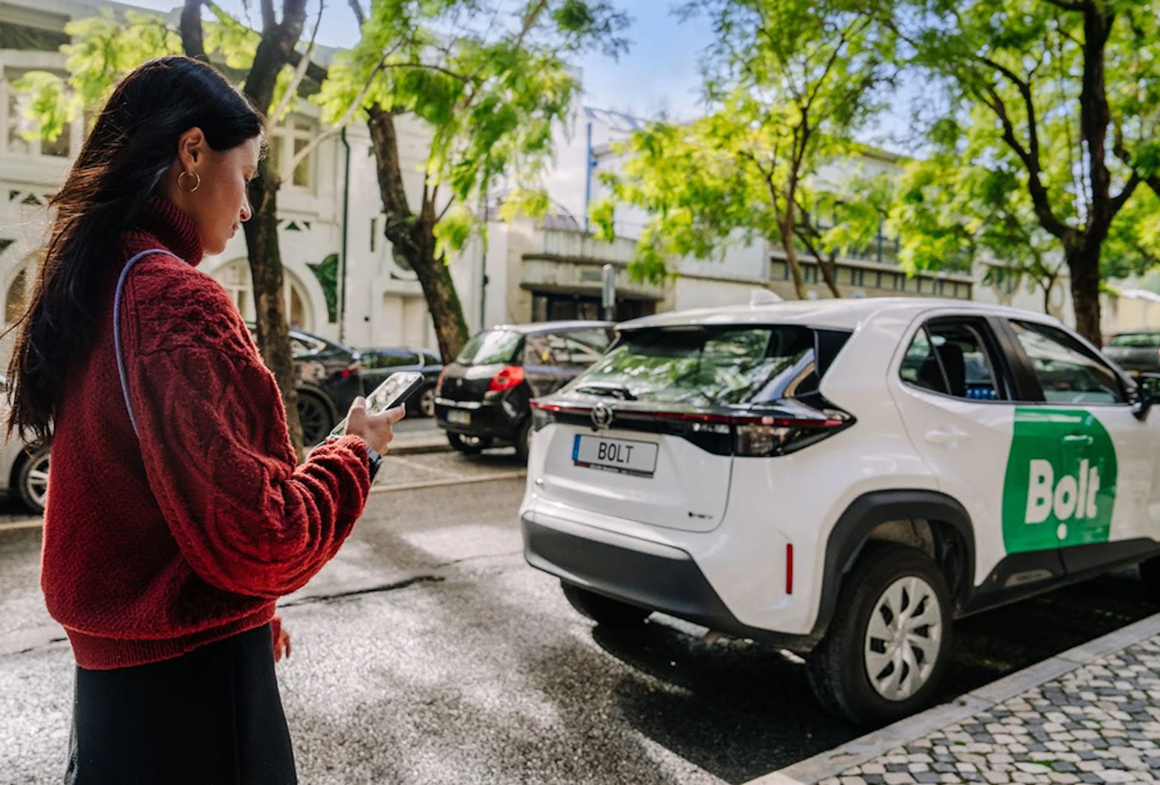 A woman in a red jumper looks at her phone as a Bolt car stops to pick her up