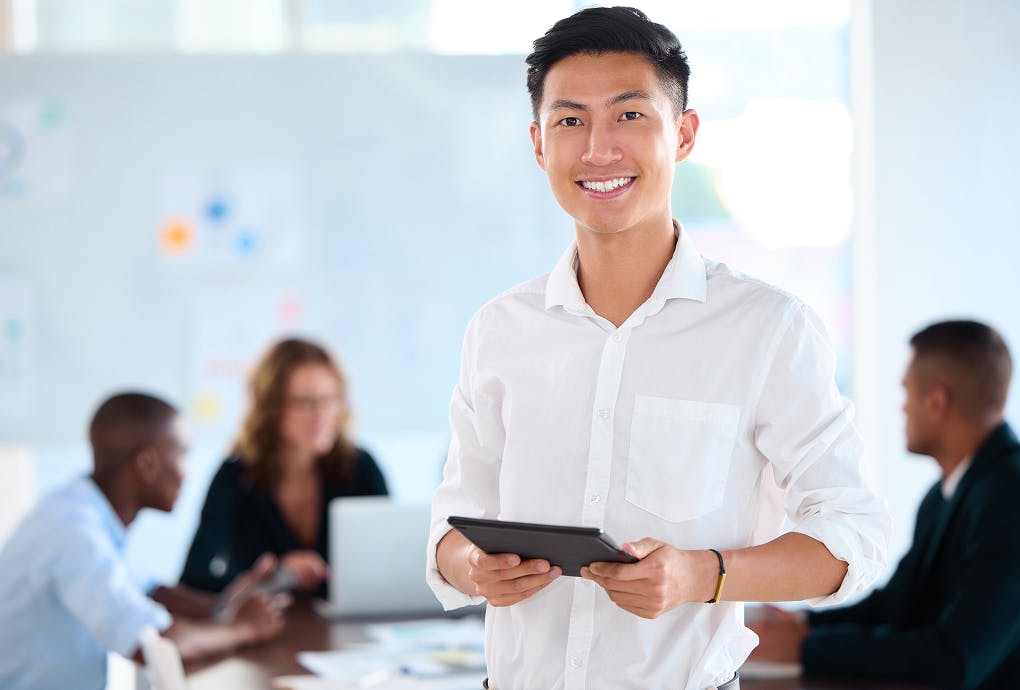 A man in a white shirt holds an ipad, looking to camera. In the background, his team members are engaged in a meeting.