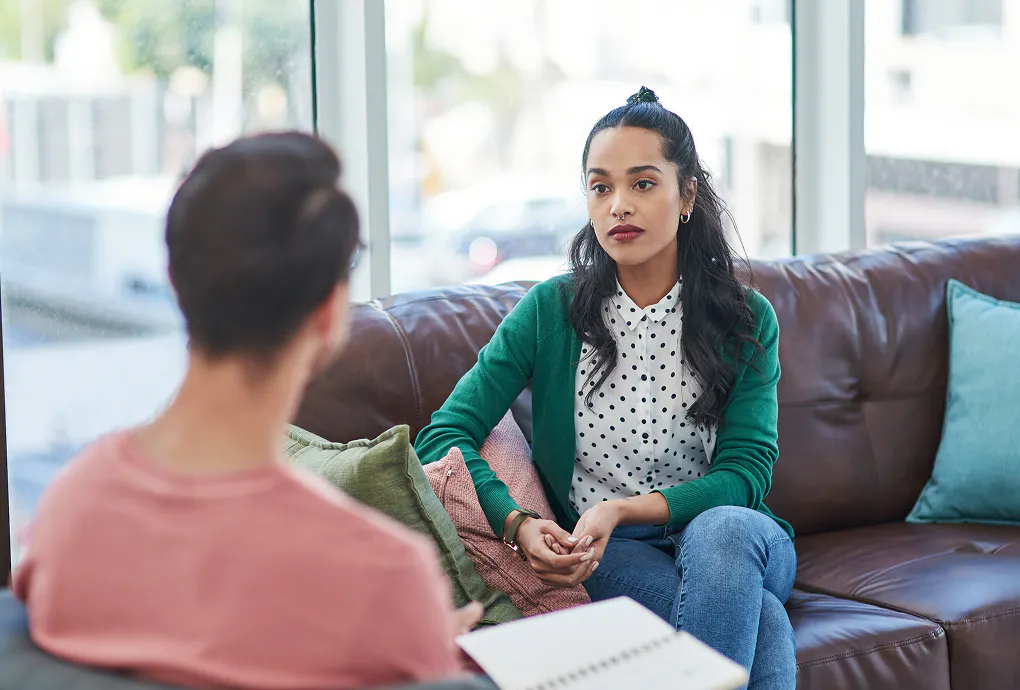 Two colleagues are having a conversation. The woman faces towards us, sat on a brown leather sofa. The man faces away from us, with only the back of his head in view.