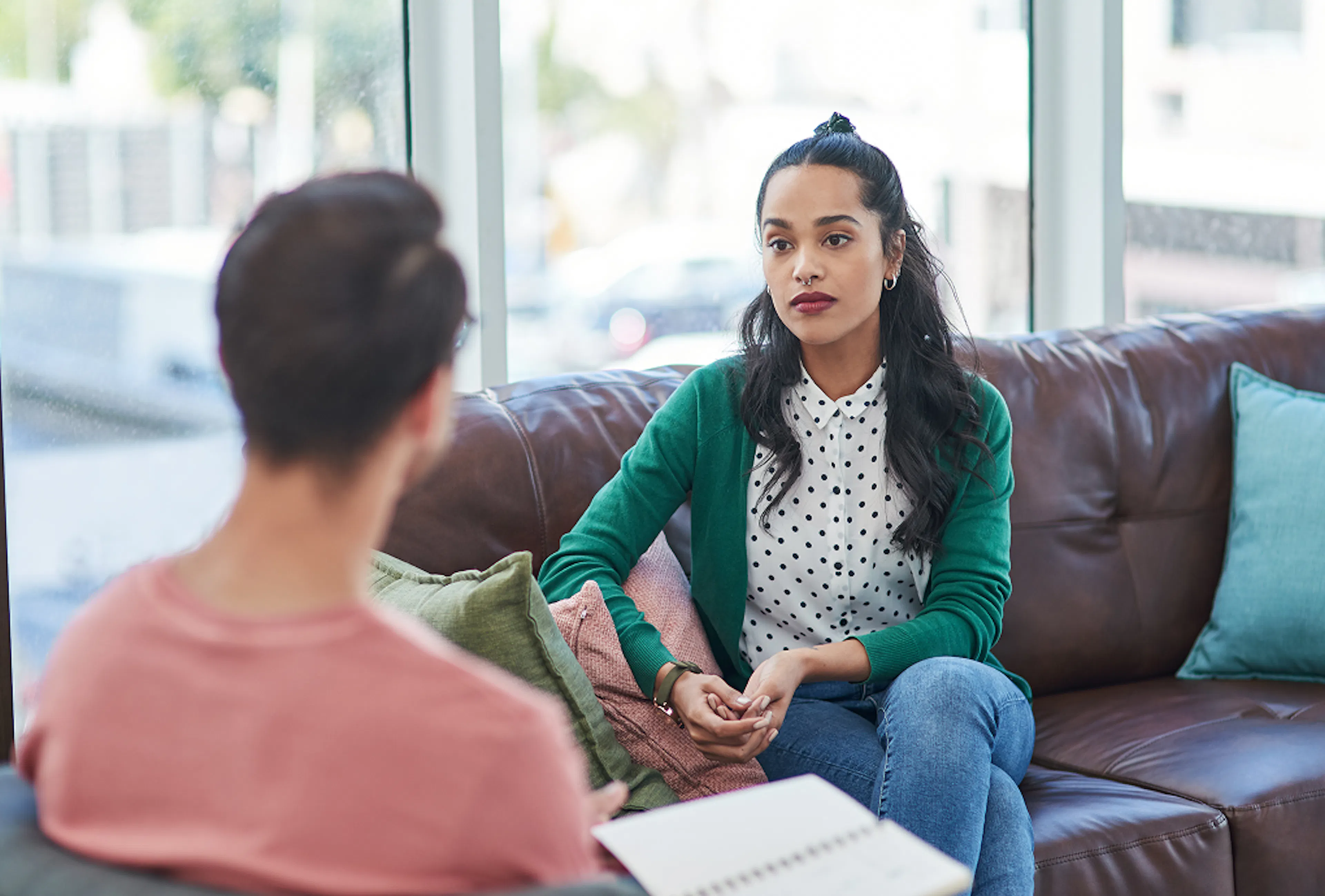 Two colleagues are having a conversation. The woman faces towards us, sat on a brown leather sofa. The man faces away from us, with only the back of his head in view.
