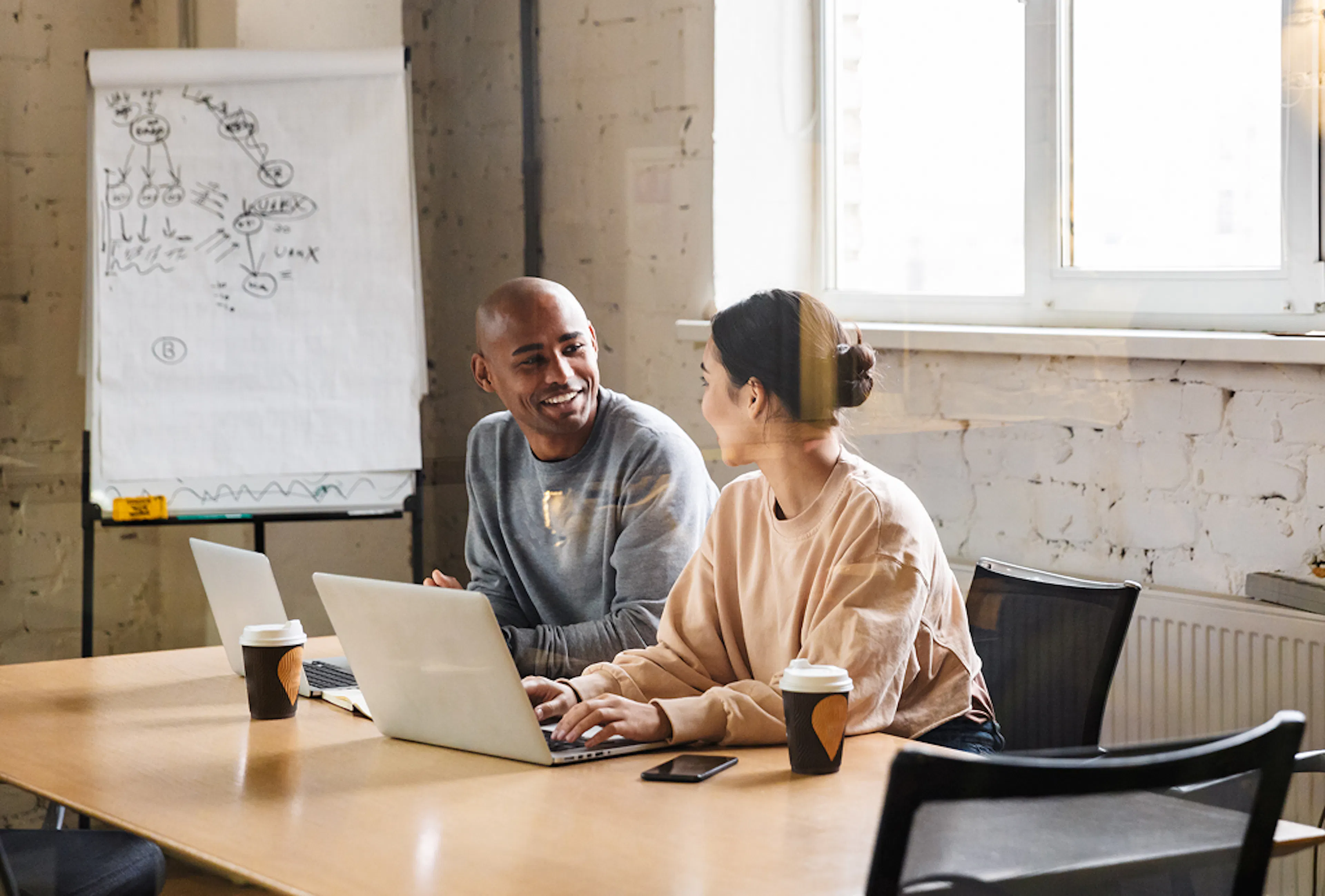 Two team members sit side by side in an office, talking to one another. Their laptops are open on the desk in front of them.