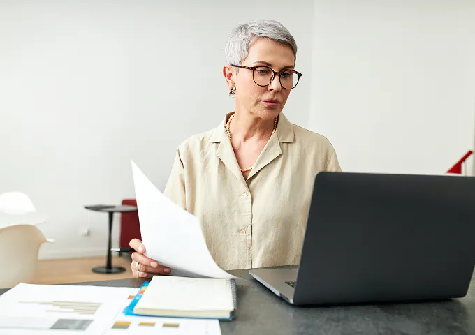 A mature business woman sits at her desk, holding papers in one hand and looking at her laptop screen in the other.