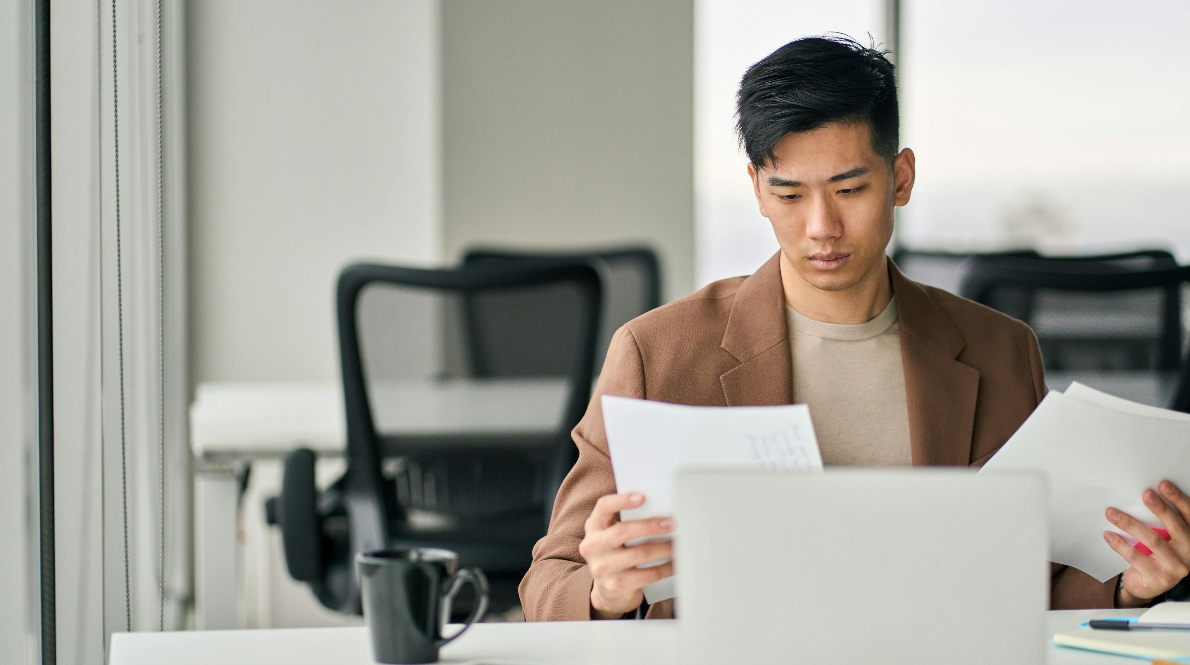 A businessman sits at his desk in front of a laptop, paper in hand.