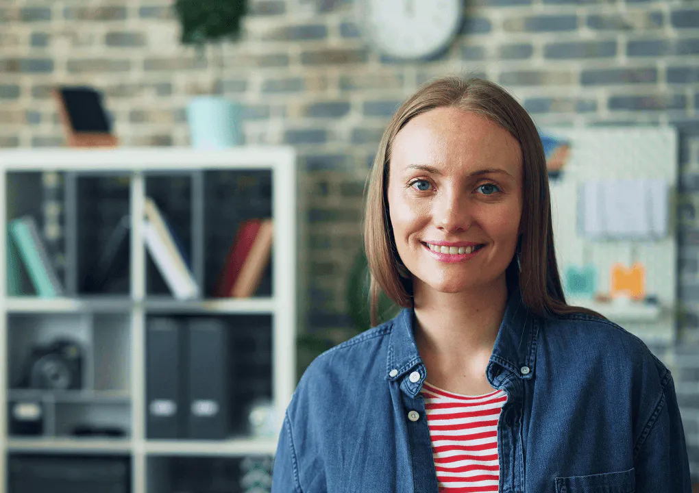 A female employee looks to camera, smiling. She's wearing a red striped top and a blue overshirt. In the background is a bookshelf containing files and tech.