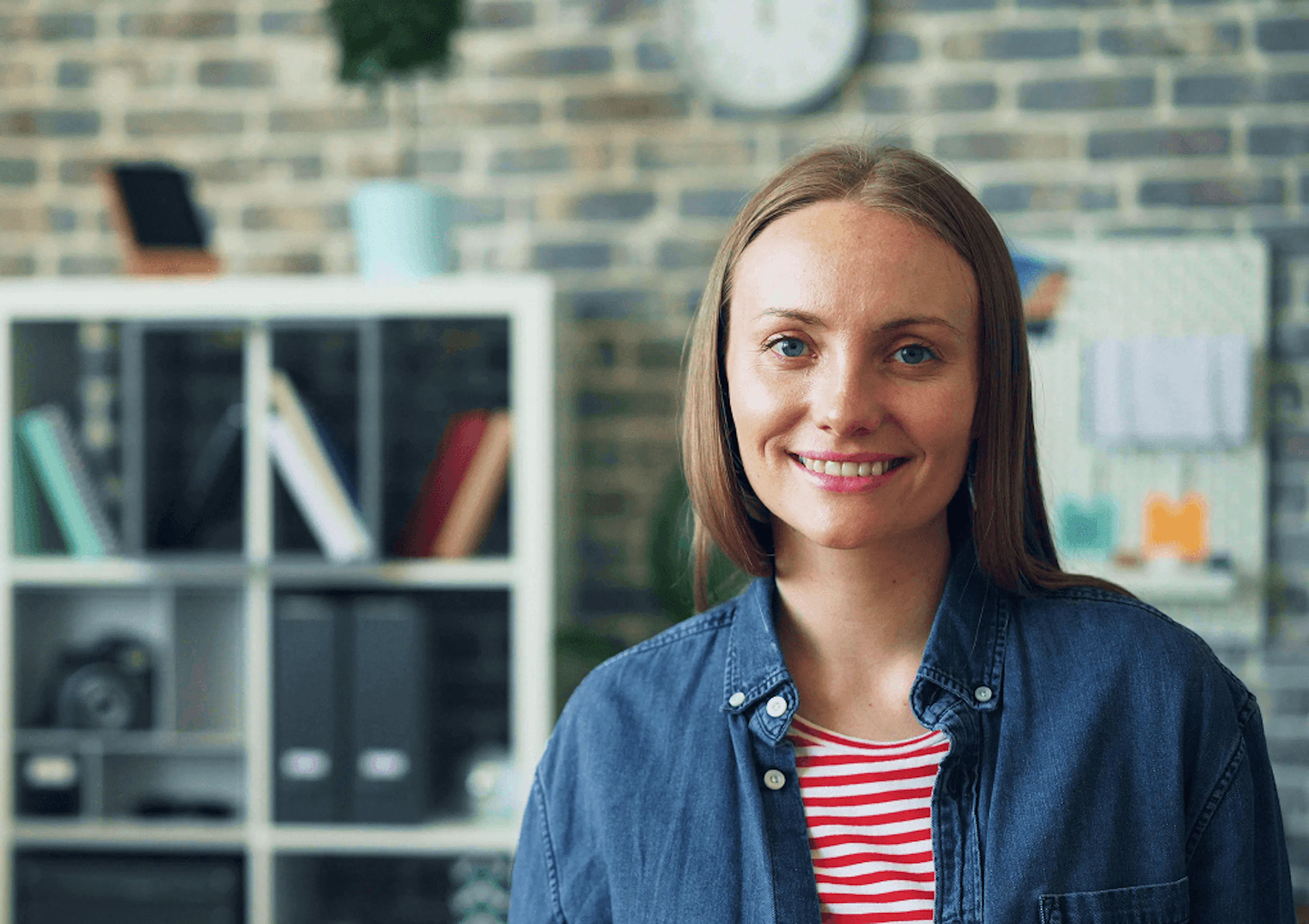 A female employee looks to camera, smiling. She's wearing a red striped top and a blue overshirt. In the background is a bookshelf containing files and tech.