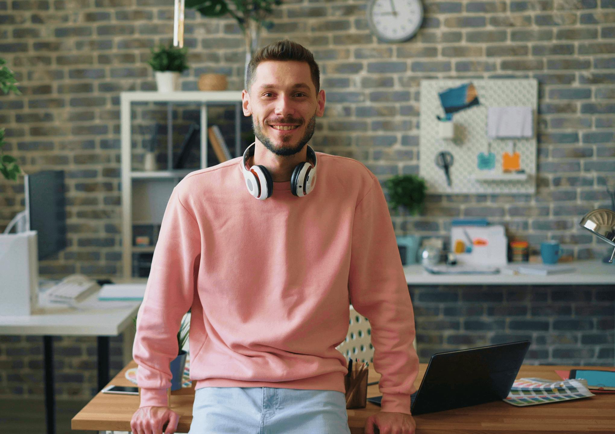 Male coworker wears a pink jumper and headphones around his neck, looking to camera
