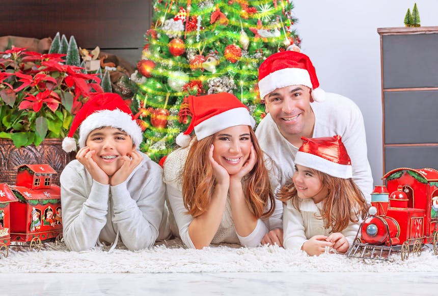 Vader, moeder en kinderen liggen voor de kerstboom met een kerstmuts op