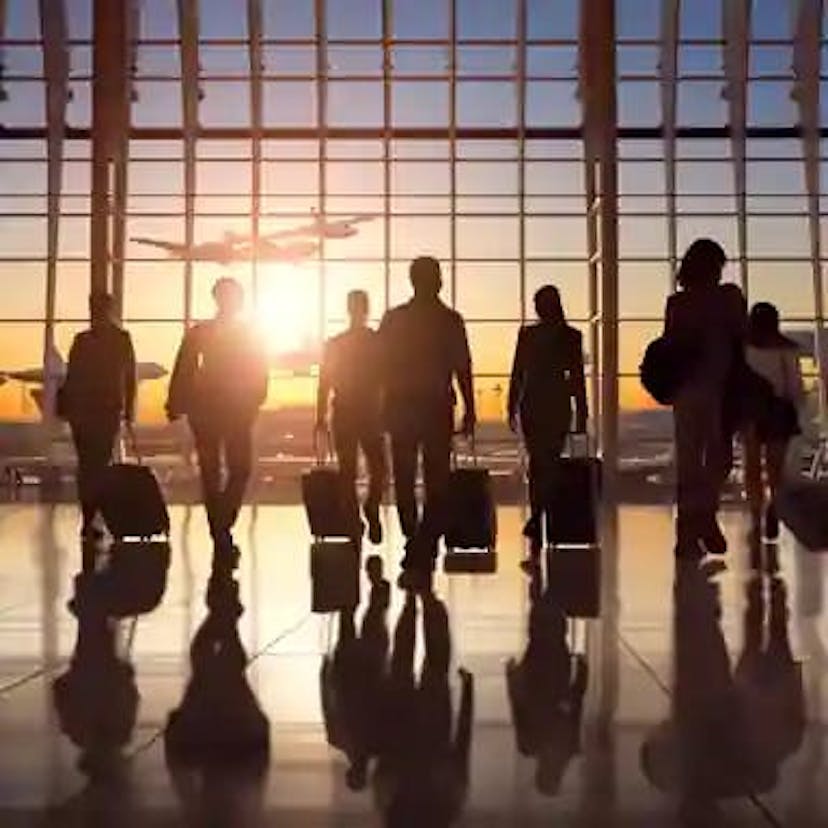 Main post image that shows a group of people walking through an airport terminal, all with rolling suitcases. In the background, through large glass windows, a sunrise or sunset is visible, with an airplane in flight seen in the distance. The image conveys the typical atmosphere of air travel, emphasizing movement and transition, captured in a moment of natural light that highlights the silhouettes of the travelers.