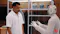 A man in a white short-sleeve shirt stands facing a white humanoid robot with glowing circular ear panels, as if in conversation, inside a wood-paneled showroom with shelves displaying knitted garments and accessories.