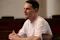 A man with short dark hair, dressed in a plain white t-shirt, sits at a wooden desk in a classroom. He appears to be engaged in conversation. The classroom is empty except for him, with several rows of wooden desks visible in the background.
