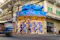 Street corner in Bangkok featuring a colorful “I Wanna Feel Thai” billboard above a building, with pedestrians, a cyclist, and a blurred vehicle passing by.