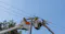 Two utility workers in bucket trucks repair power lines on a wooden pole under a clear blue sky.