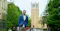 A man wearing a blue blazer and white shirt stands outdoors on a university campus. Behind him is a large clock tower made of light brown brick, surrounded by green trees and other campus buildings. The sky is clear, and a few people are walking in the background.