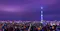 A panoramic nighttime cityscape with a purple sky, showing a vast expanse of illuminated buildings. The Tokyo Skytree, a tall, slender tower glowing with blue and white lights, stands prominently in the center against the skyline.