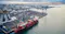 Aerial view of a cargo ship docked at a container port beside a coastal city under cloudy skies.