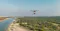 The image shows a drone flying over a coastal landscape with a river or inlet on the left and a dense forest on the right. The scene is expansive, with a clear sky and distant landforms visible in the background. The drone appears to be equipped with a camera or sensor, suggesting it might be used for aerial photography, surveying, or environmental monitoring.