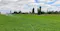 The image shows a lush green field being irrigated with a sprinkler system. The sprinklers are evenly spaced, watering the crops under a partly cloudy sky. Tall trees line the background, and the landscape appears to be flat and expansive.