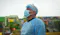The image shows a person wearing a blue protective gown, a hairnet, and a face mask, with glasses resting on top of the hairnet. The background features colorful structures and greenery, suggesting an outdoor setting, possibly in a community or residential area.
