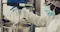The image shows a person in protective clothing, including a mask, goggles, and gloves, working in a laboratory or industrial setting. They are pouring a substance from a container into a large mixing vessel, which is likely part of a manufacturing or research process. The environment suggests a focus on safety and precision, possibly in the pharmaceutical or chemical industry.