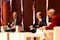 The image depicts three individuals engaged in a panel discussion or conference setting. They are seated on stage, each holding papers, and one person is speaking into a microphone. The setting suggests a formal event or meeting.