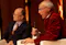 The image shows two individuals seated in a formal setting, with one person holding a microphone, suggesting a panel discussion or interview. The individual with the microphone is wearing a red jacket.