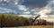 The image depicts a scene of agricultural activity, specifically the harvesting of sugarcane. It shows machinery at work, with a tractor and harvester operating in a field of tall sugarcane plants. The sun is setting or rising in the background, casting a warm glow over the scene, and the sky is filled with clouds, adding a dramatic effect to the landscape. This image highlights the mechanized process of sugarcane harvesting, which is an essential part of agricultural production.