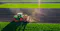 The image depicts a tractor working on a farm, likely involved in planting or cultivating crops. The scene shows a vibrant green field with neatly organized rows, indicating active agricultural activity. The tractor is equipped with machinery that appears to be used for sowing seeds or preparing the soil, highlighting the mechanized aspect of modern farming. Sunlight filters through the scene, suggesting a clear day conducive to farming operations.