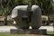 A modern abstract sculpture made of stone, featuring two large, smooth, rounded forms connected by rectangular blocks. The sculpture is set outdoors on a stone base, surrounded by grass and trees, suggesting it is part of a public art installation in a park or garden setting. The natural backdrop enhances the sculpture's aesthetic, highlighting the contrast between the organic environment and the geometric, polished artwork.